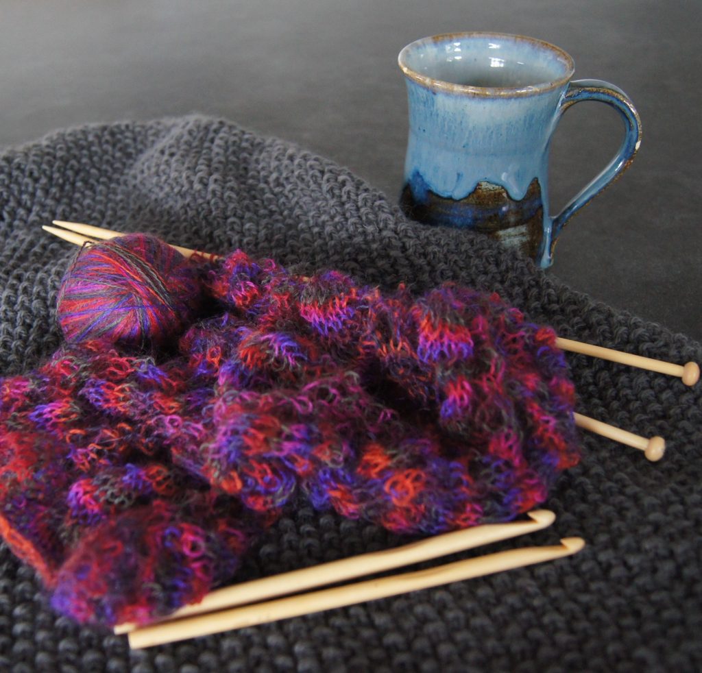 Photo of knitting, yarn, crochet hooks, and a pottery mug